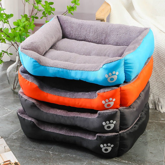 Stack of rectangular dog beds in blue, orange, and black with soft gray interior and paw print logo, displayed in a cozy indoor setting
